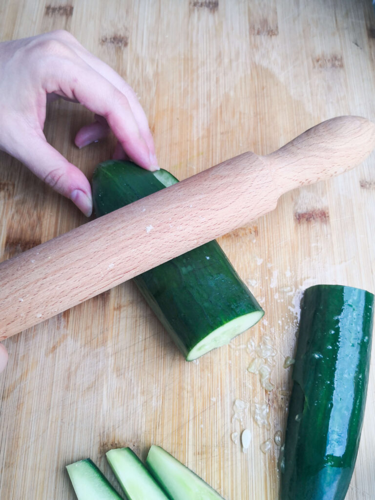 Smacking a chopped cucumber with a large wooden rolling pin on a wooden board.