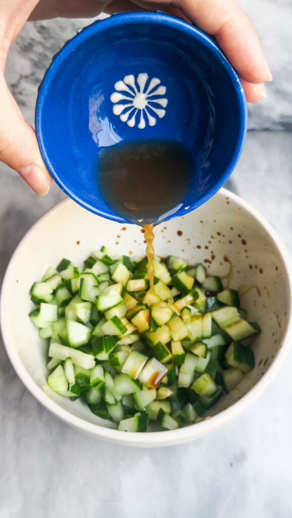 Soy sauce being poured into a small white bowl of diced cucumber on a grey marble board.
