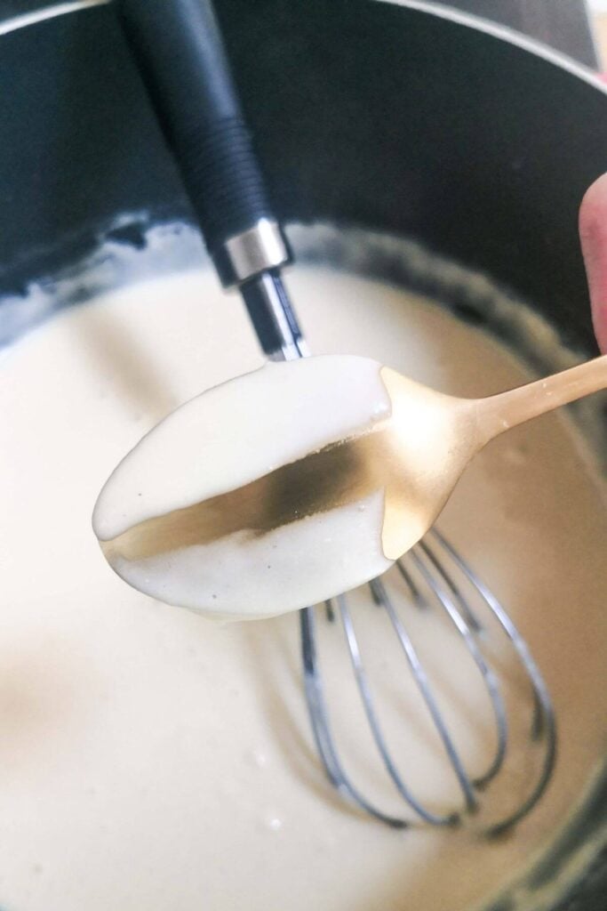 A small gold spoon with cheesy white sauce on it, with a pot of sauce in the background.