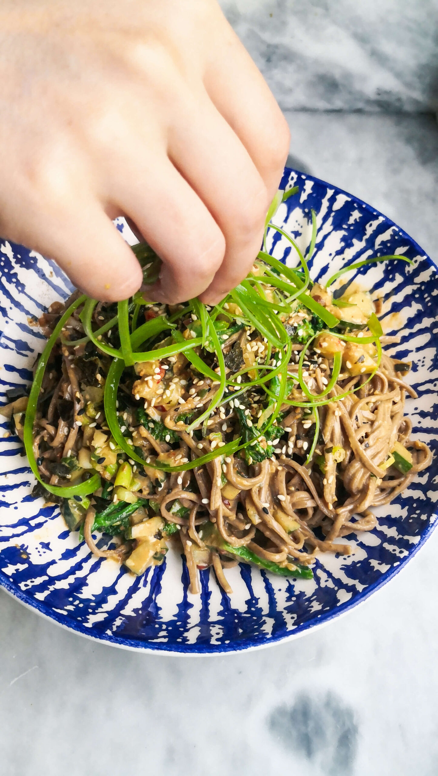Hand placing curly spring onions on top of a bowl of sesame soba noodles.