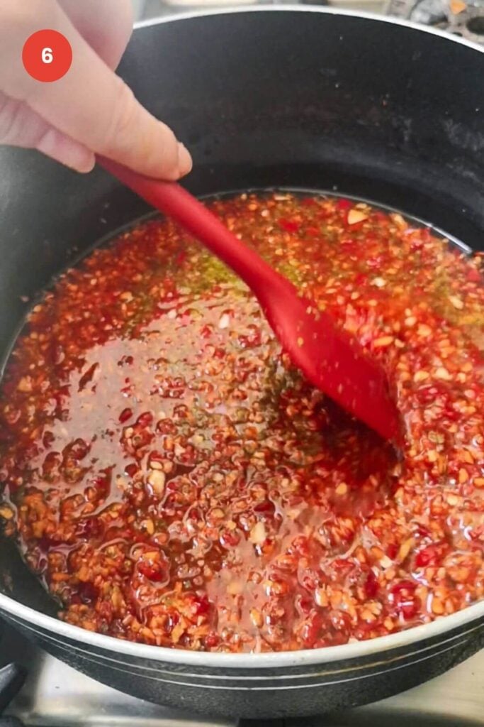 Red spatula stirring chili jam in a large pot.