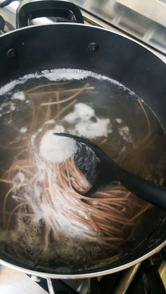Soba noodles cooking in a large black pot, with a large black spoon stirring them.