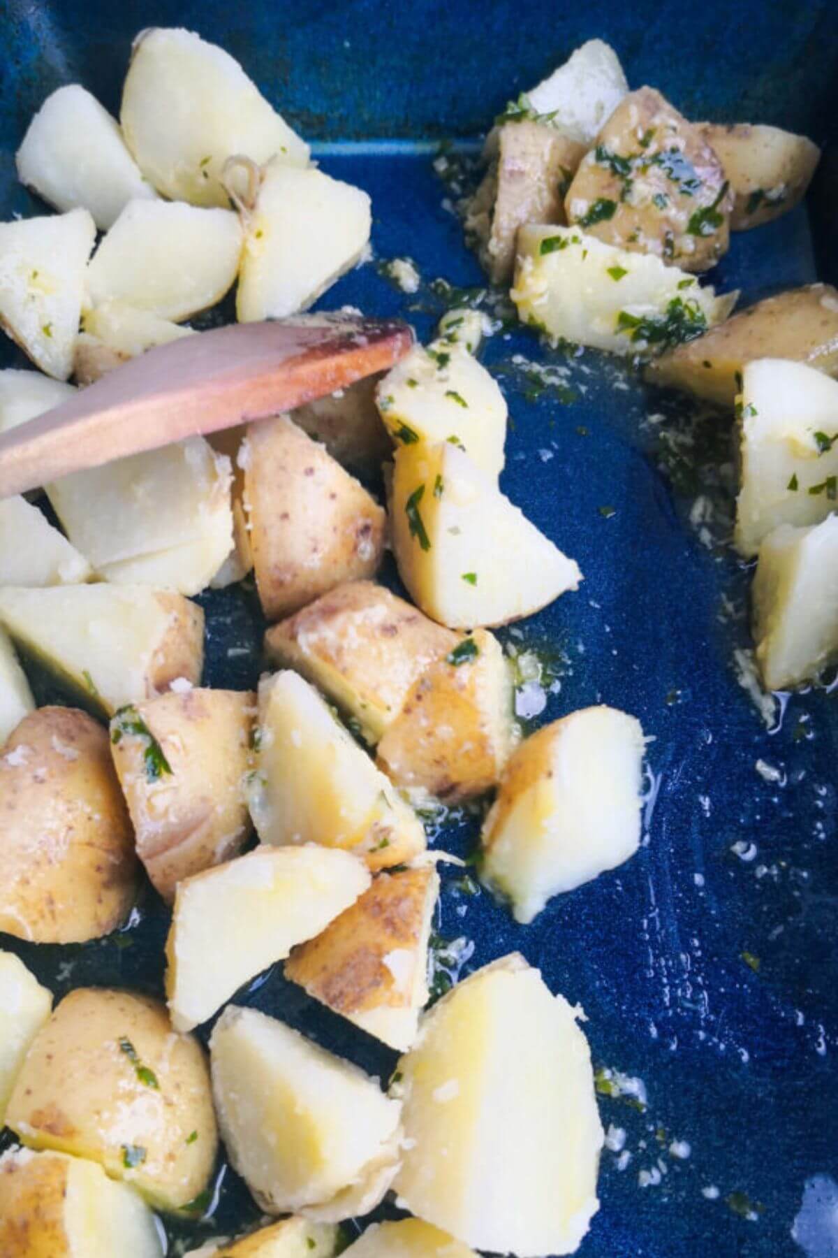 Garlic butter sauce being mixed through potato chunks in a large blue oven dish with a wooden spoon.