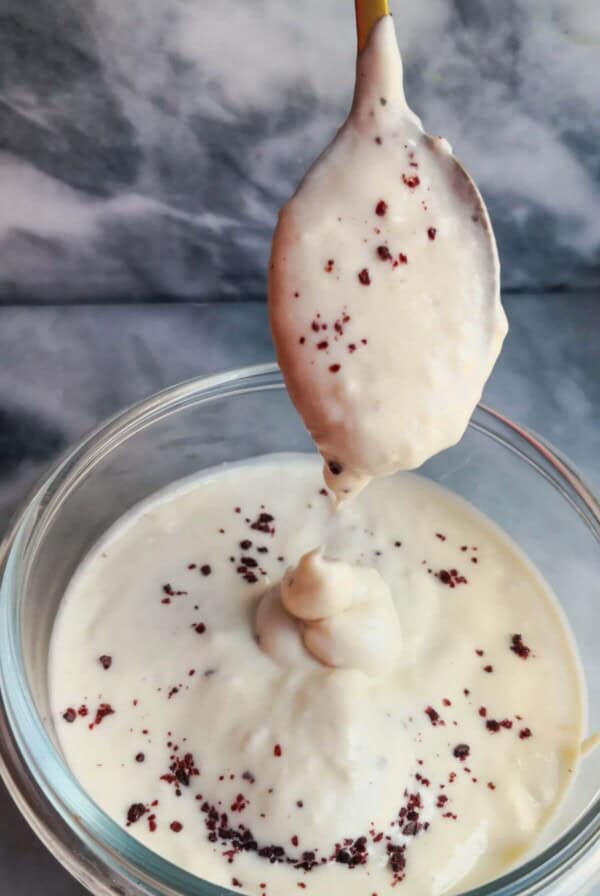 Tahini yogurt sauce being poured off a small gold spoon into a small glass bowl on a grey marble background.