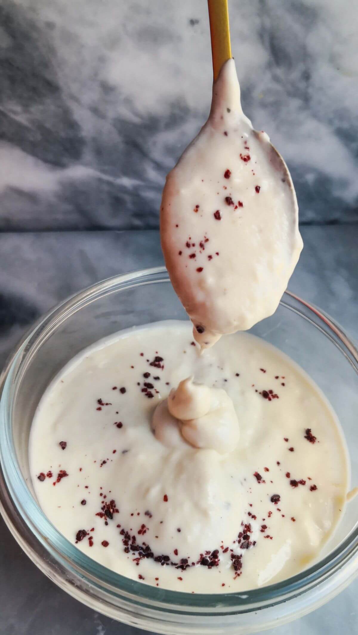 Tahini yogurt sauce being poured off a small gold spoon into a small glass bowl on a grey marble background.