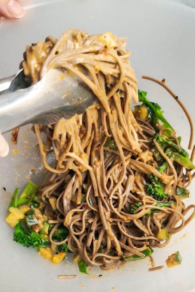 Tongs tossing soba noodles with tahini sauce.