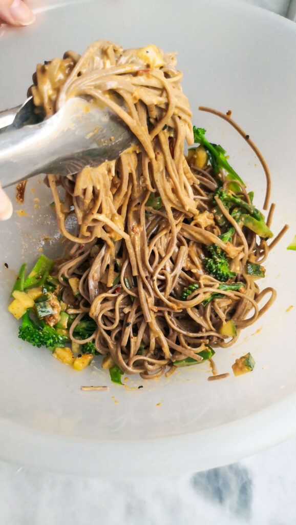 Tossing sesame soba noodle salad in a large mixing bowl with silver tongs.
