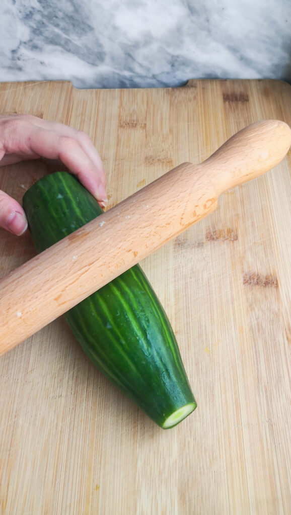 A hand holding half a cucumber and a wooden rolling pin pressing down on it.