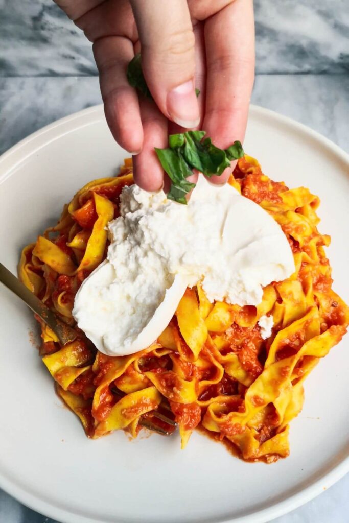 A hand adding basil to burrata on top of nduja pasta on a large white plate.
