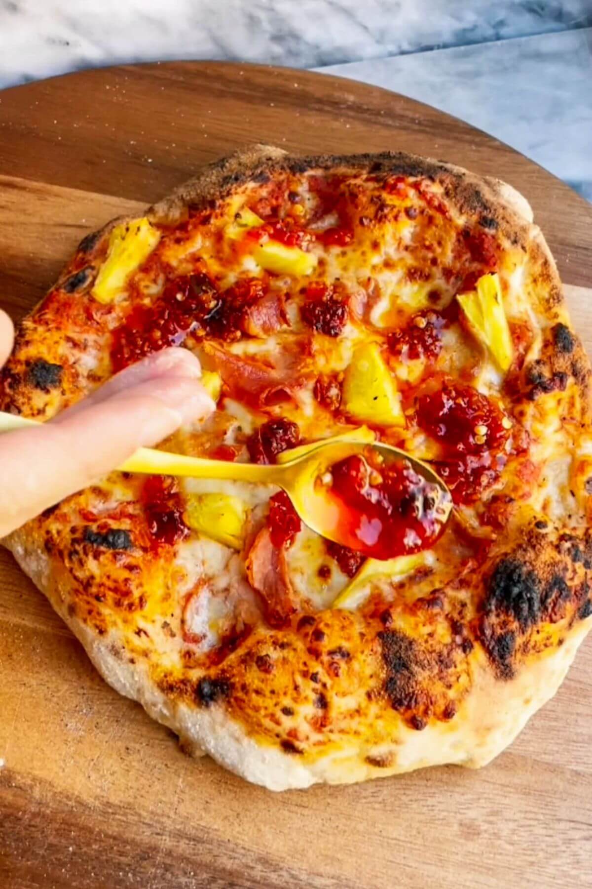 Hand holding a spoon drizzling chilli jam onto Hawaiian pizza.