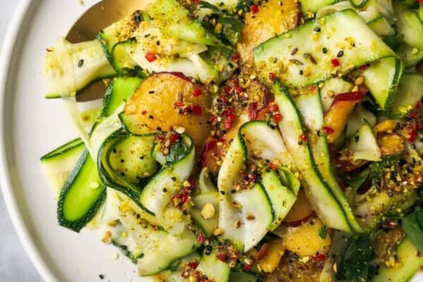 Raw courgette and peach salad on a white serving plate on a grey marble background with a small bowl of dukkah on the side.