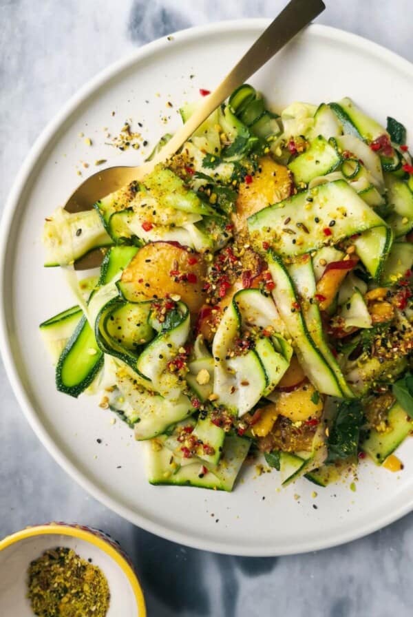 Raw courgette and peach salad on a white serving plate on a grey marble background with a small bowl of dukkah on the side.