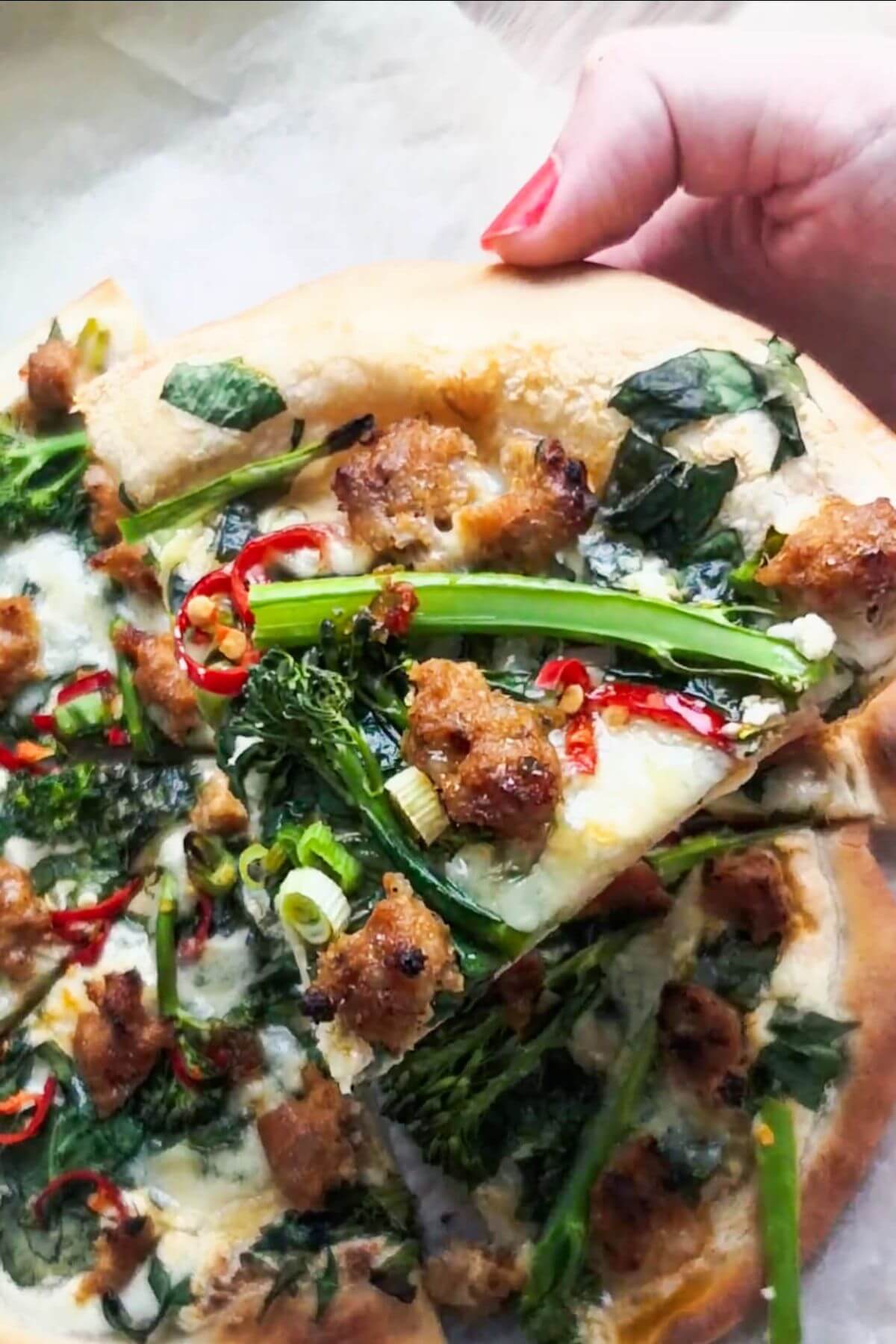 Hand holding up a slice of sausage, chilli, broccolini pizza.