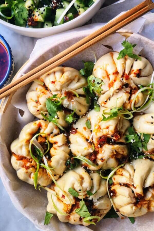 Sticky Korean chicken steamed buns in a bamboo steamer, with cucumber salad and chilli oil on the side.