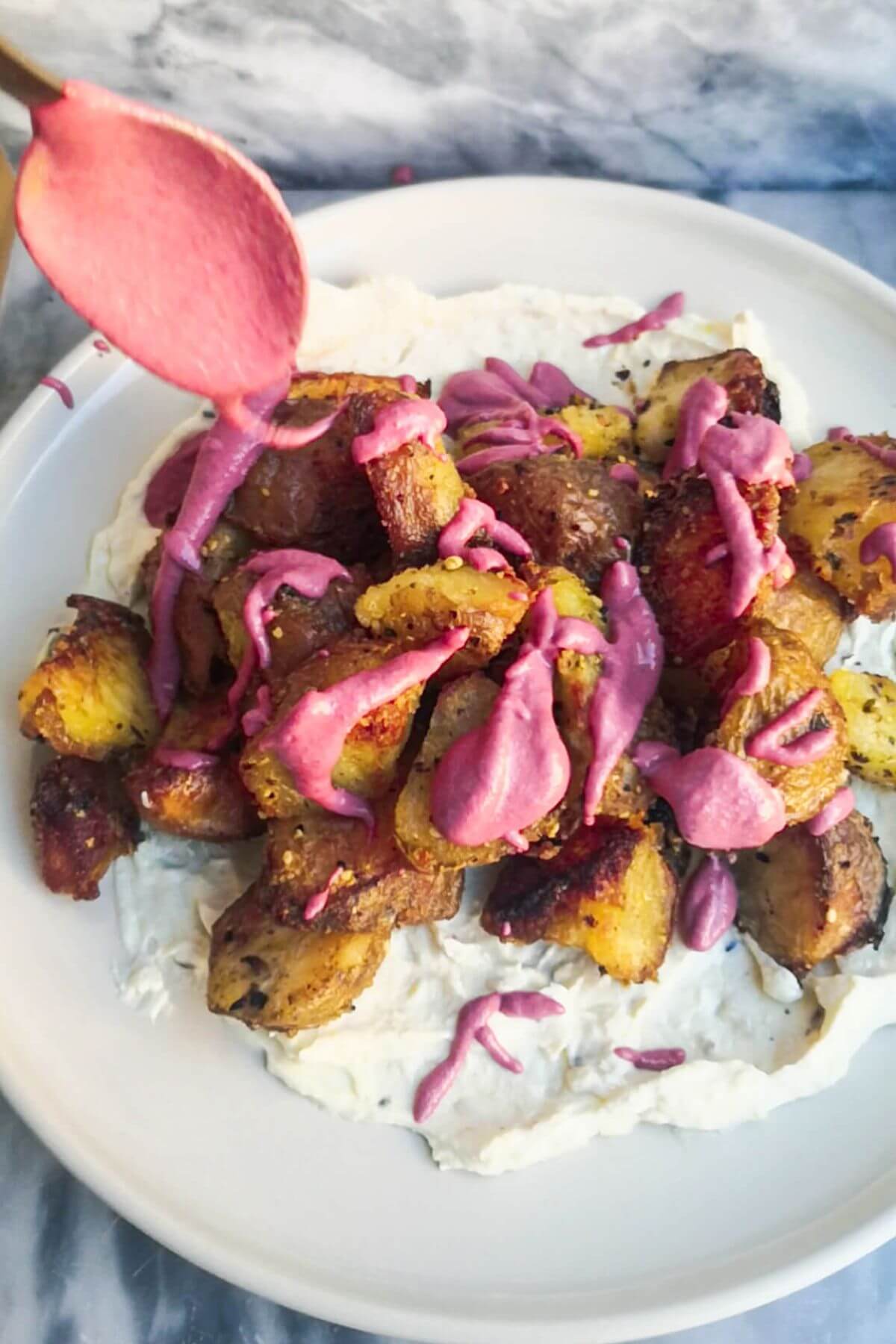 A spoon drizzling pink tahini sauce over crispy dukkah potatoes on a plate with aioli.