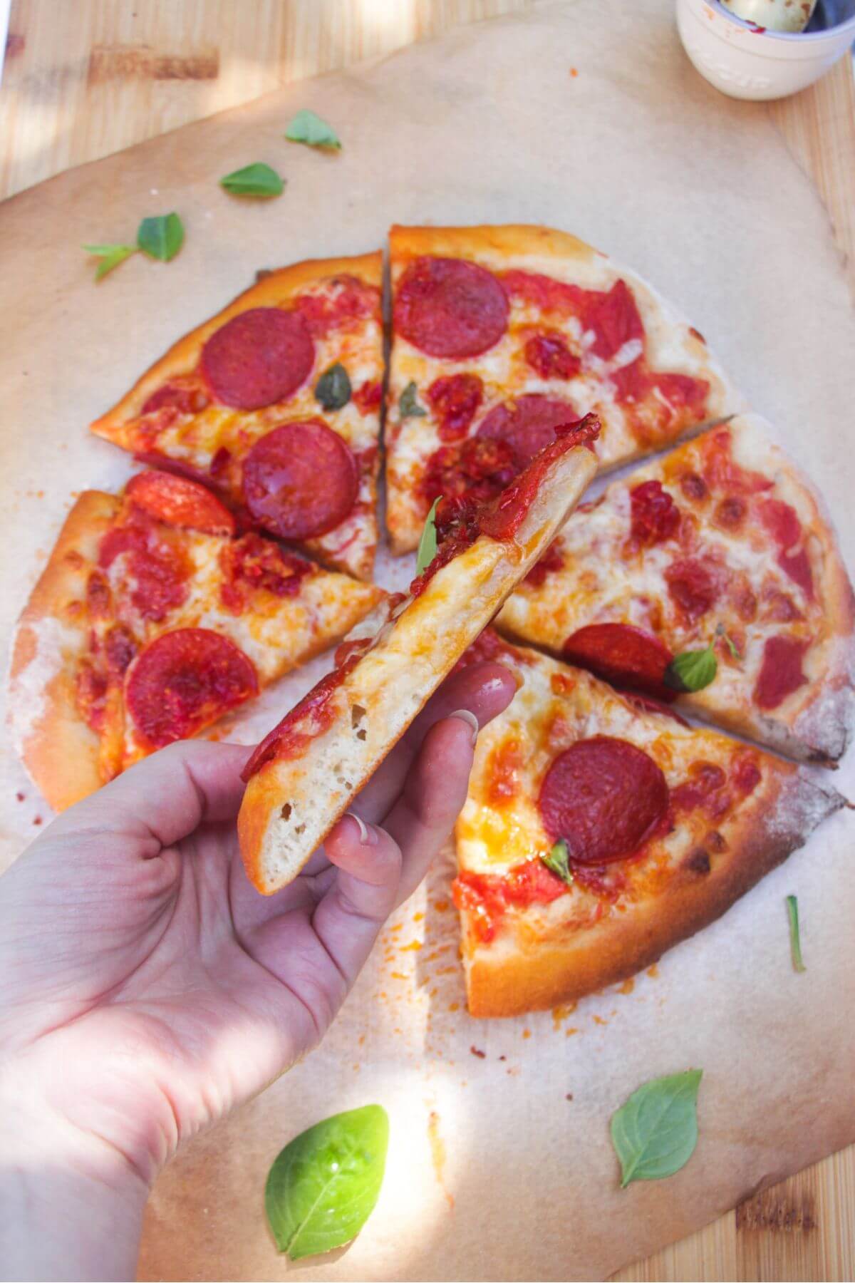 Hand holding up a slice of pepperoni pizza with the rest of the pizza in the background.