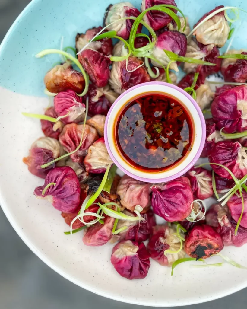 Pink and white mini dumplings in a white and blue bowl with small bowl of chilli oil in the middle.