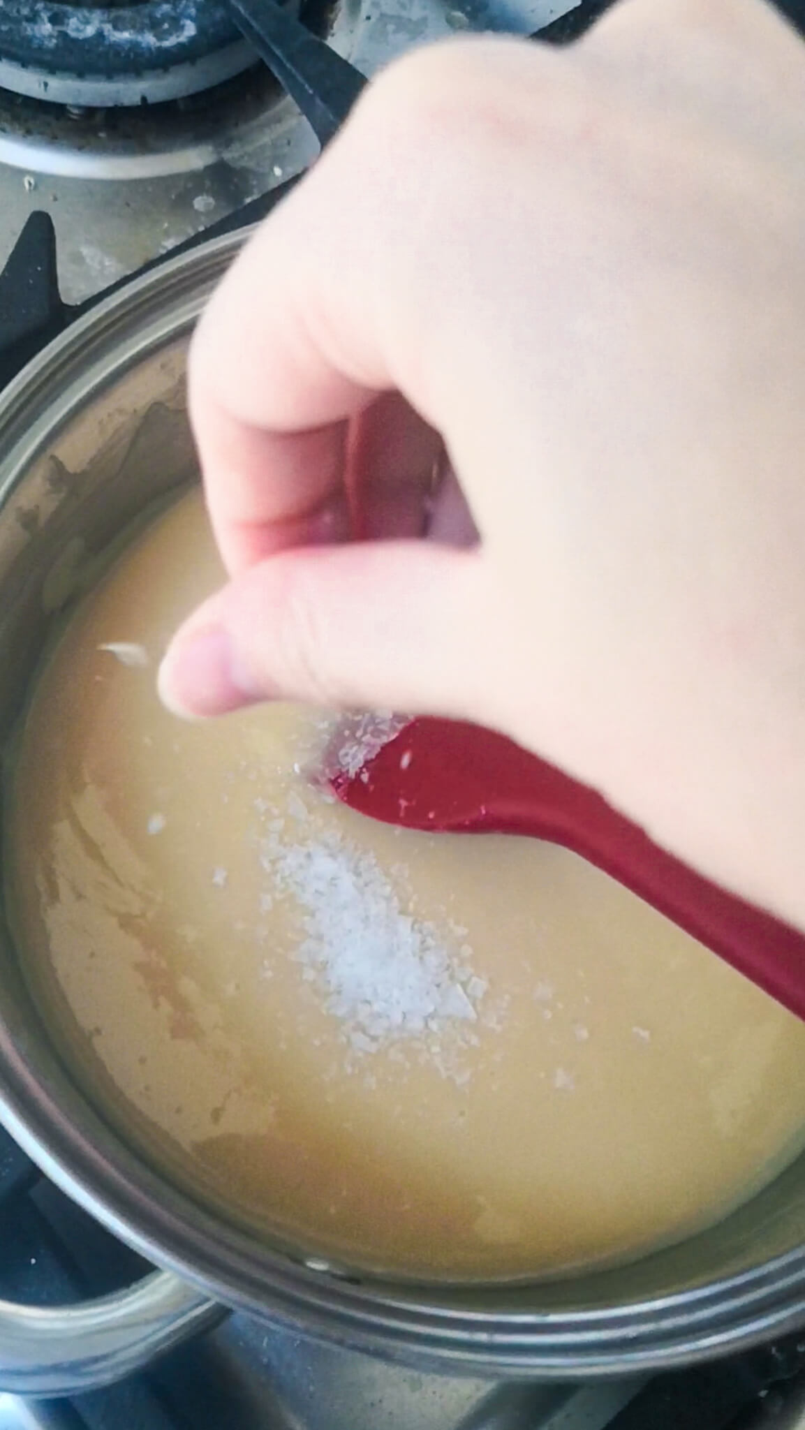 Hands adding flaky sea salt to a pot with caramel in it.
