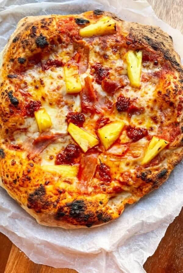 Spicy Hawaiian pizza on baking paper on a round wooden board.