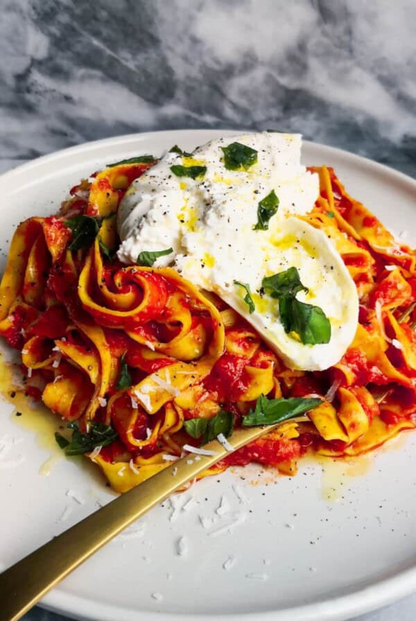 Close up view of spicy nduja pasta on a white plate with a gold fork on a grey marble background.