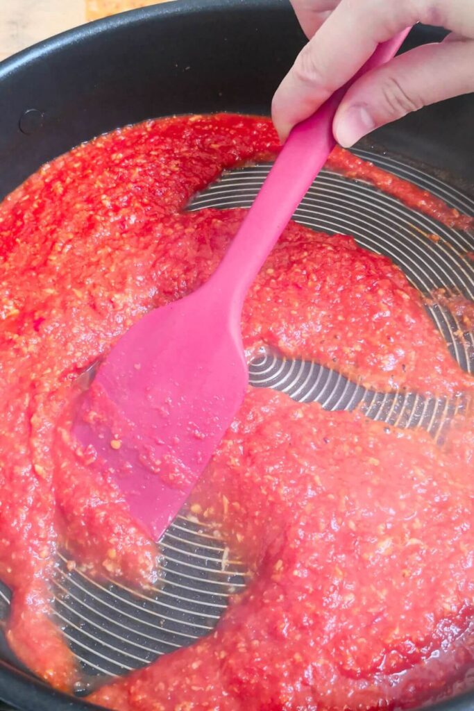 Red spatula stirring nduja tomato sauce in a large black pan.