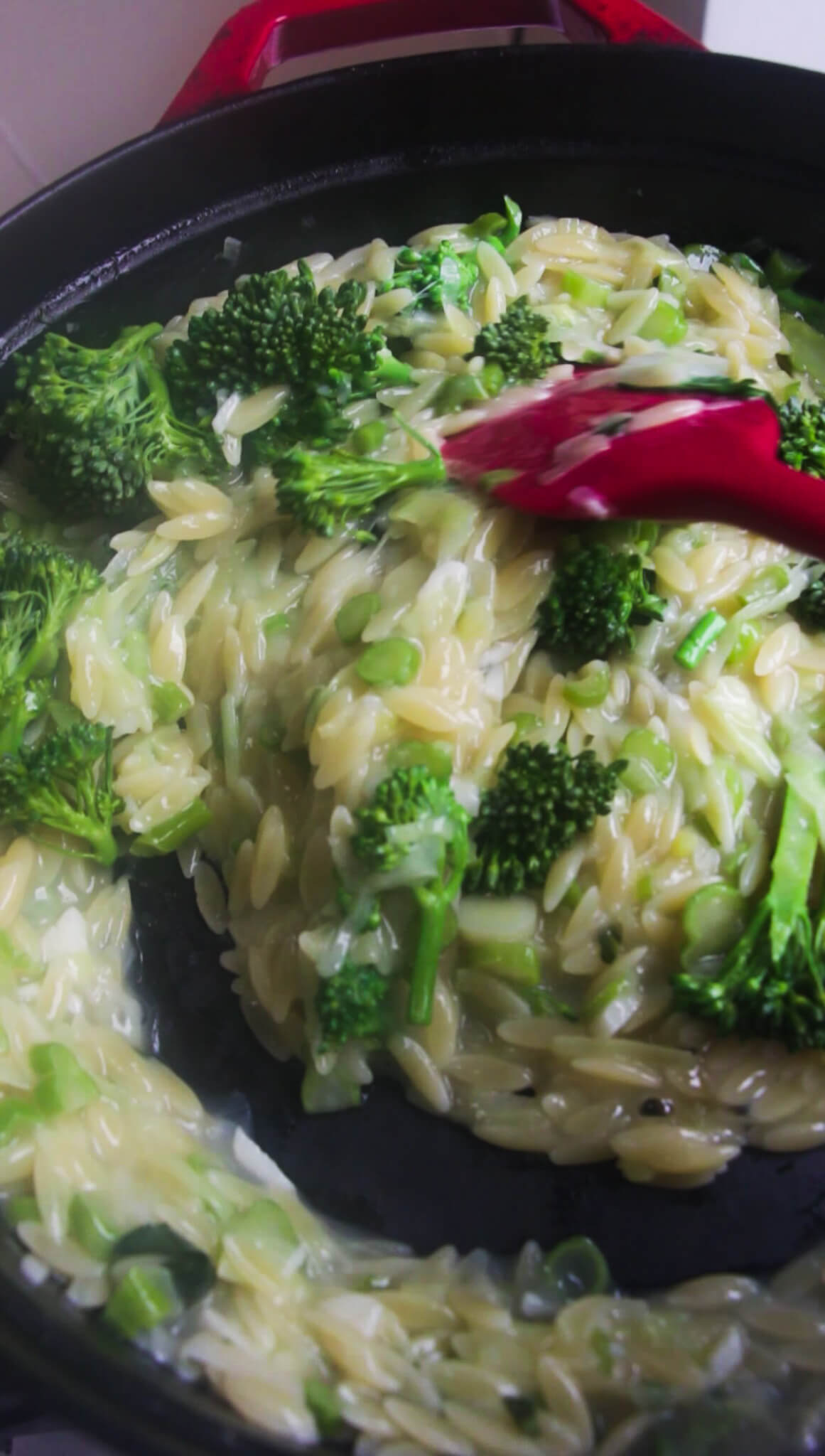 Red spatula folding broccolini into creamy orzo in a red and black pan.