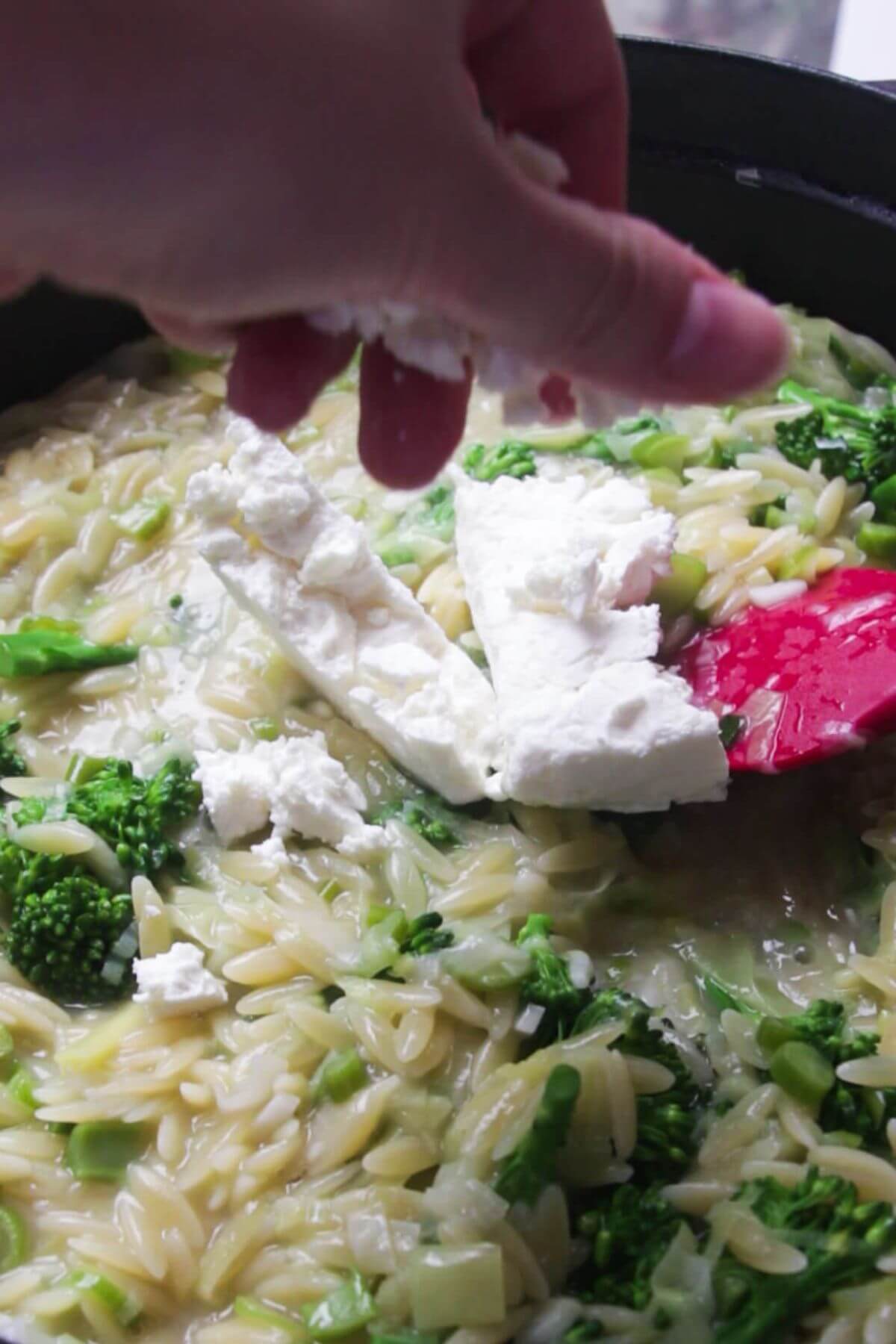Hand crumbling feta into creamy orzo in a black and red pan.