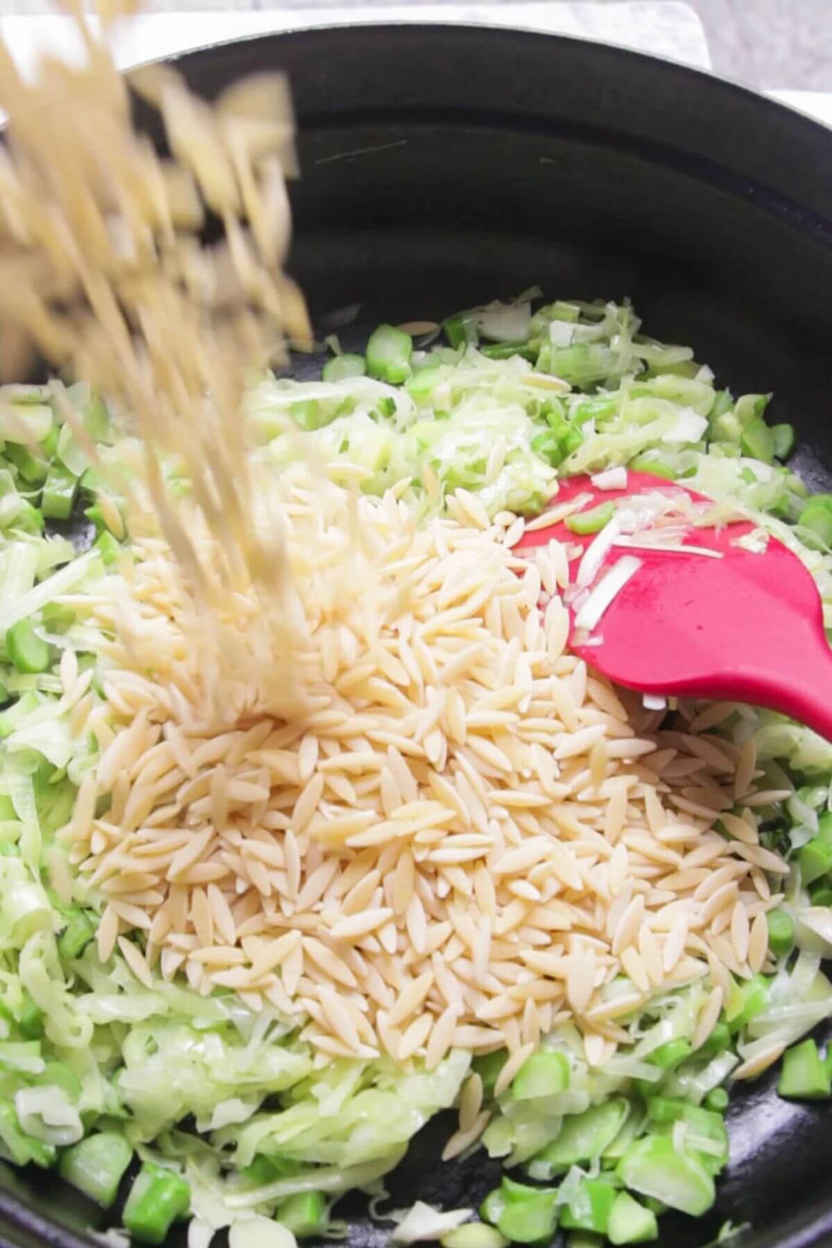 Orzo being poured into a black and red pan with leeks, broccolini and garlic.