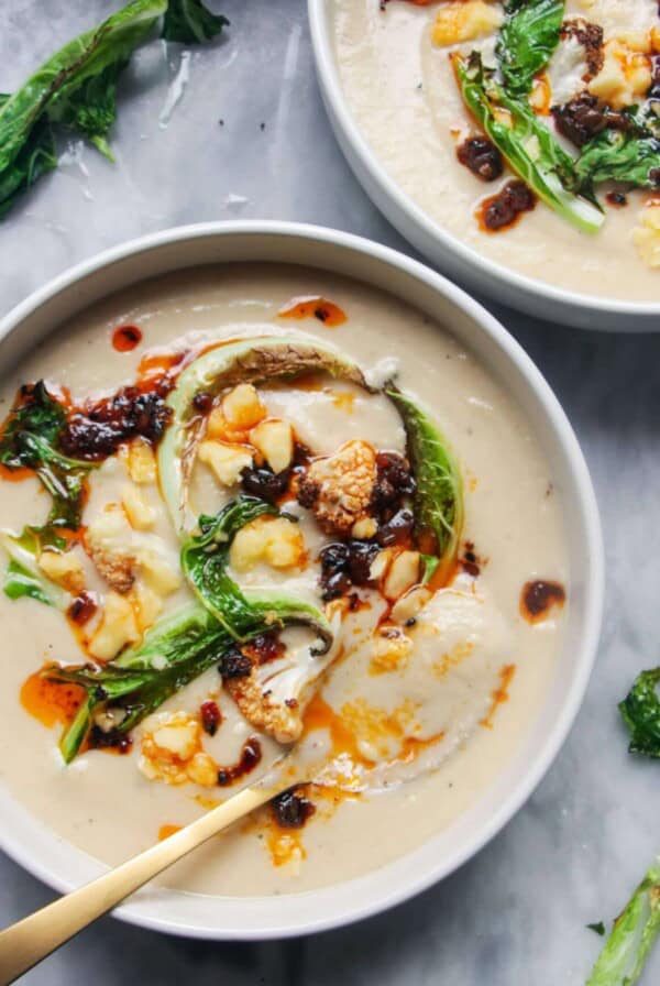 Close up of roasted cauliflower cheese soup in a bowl with another bowl just out of frame, on a grey marble background.