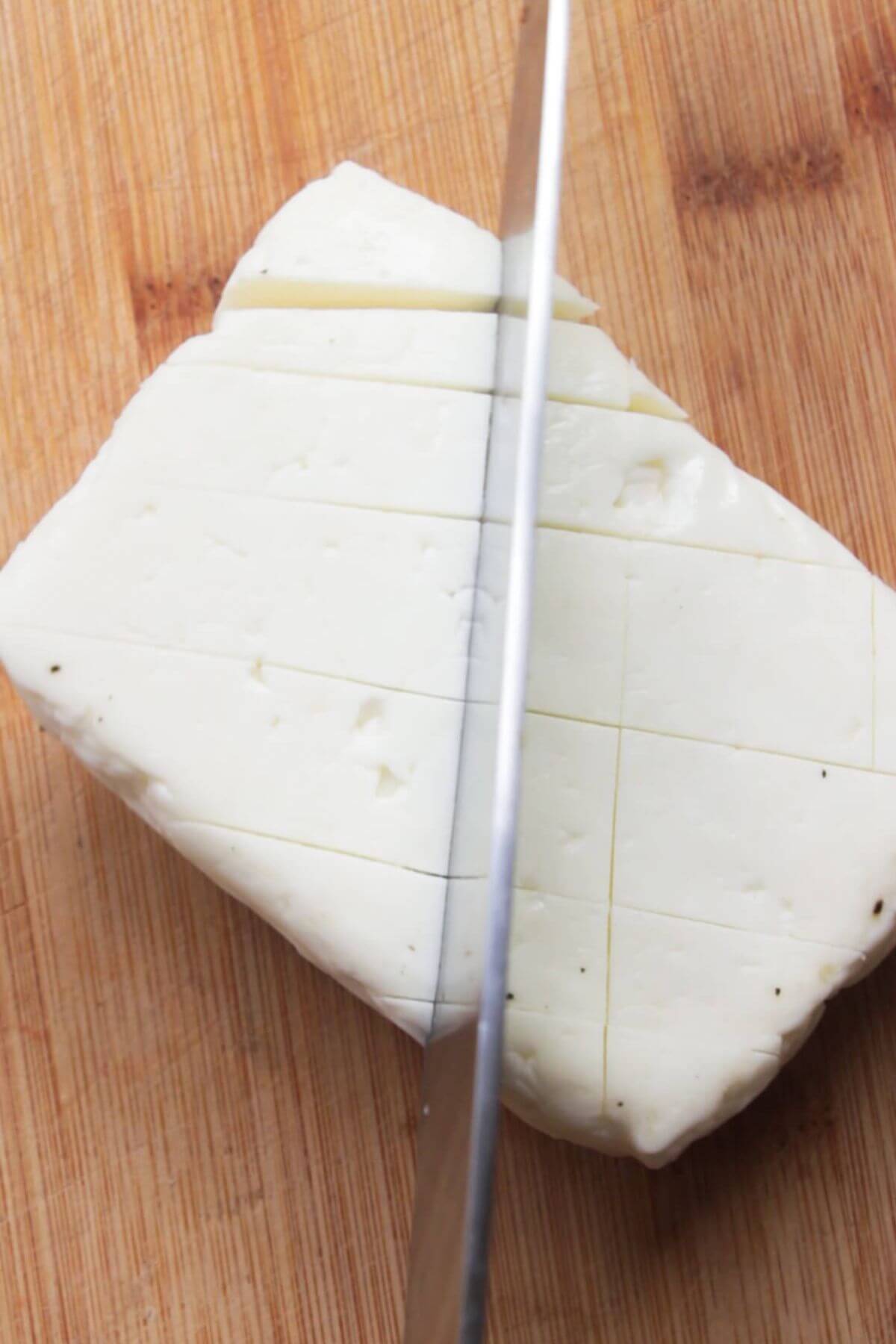 A knife cutting criss cross lines into a whole block of halloumi on a wooden board.