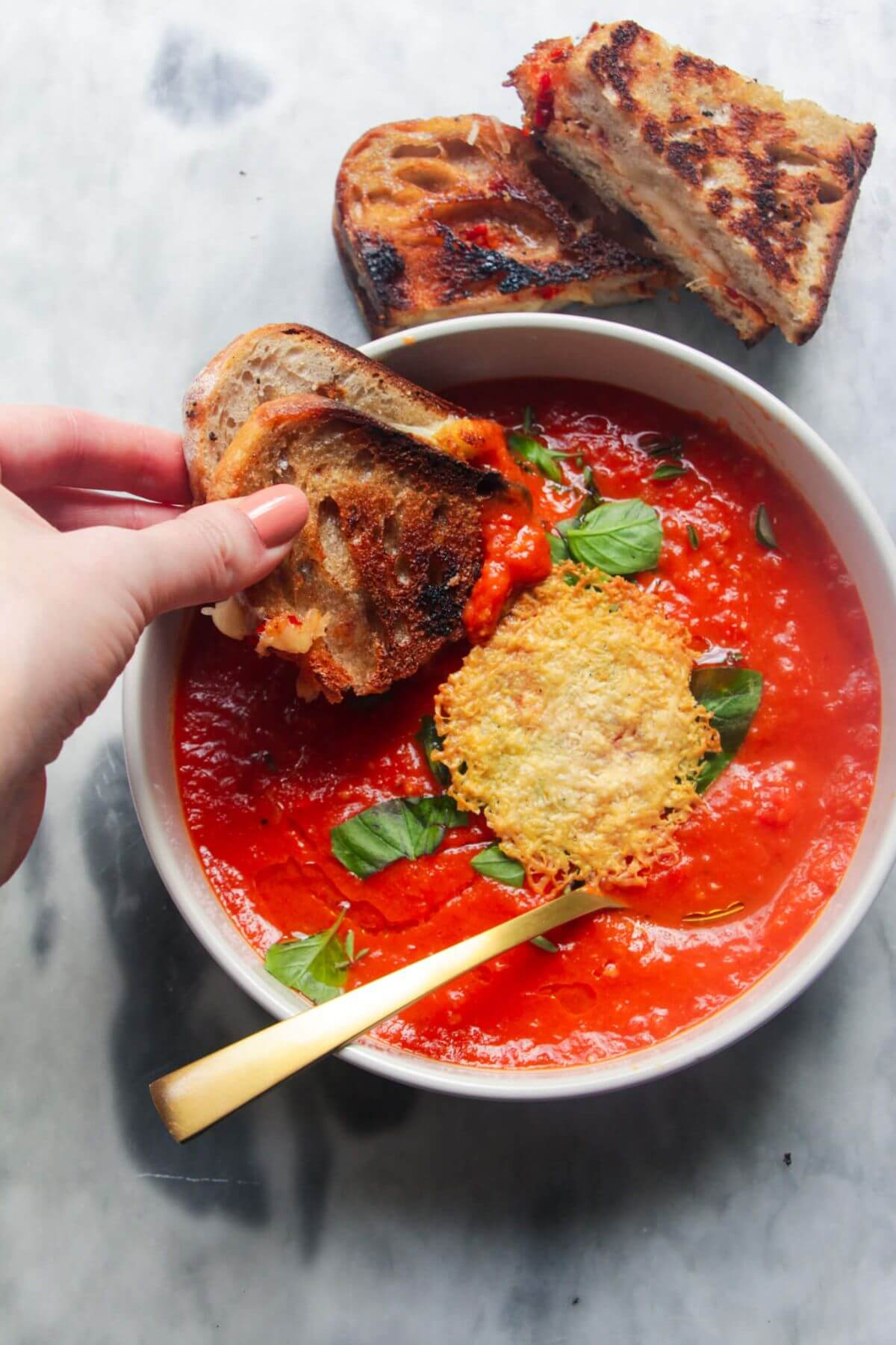 Hand dipping grilled cheese into tomato and red pepper soup on a grey marble background.