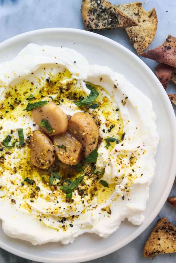 Labneh with confit garlic and dukkah on a plate with pita chips on the side.