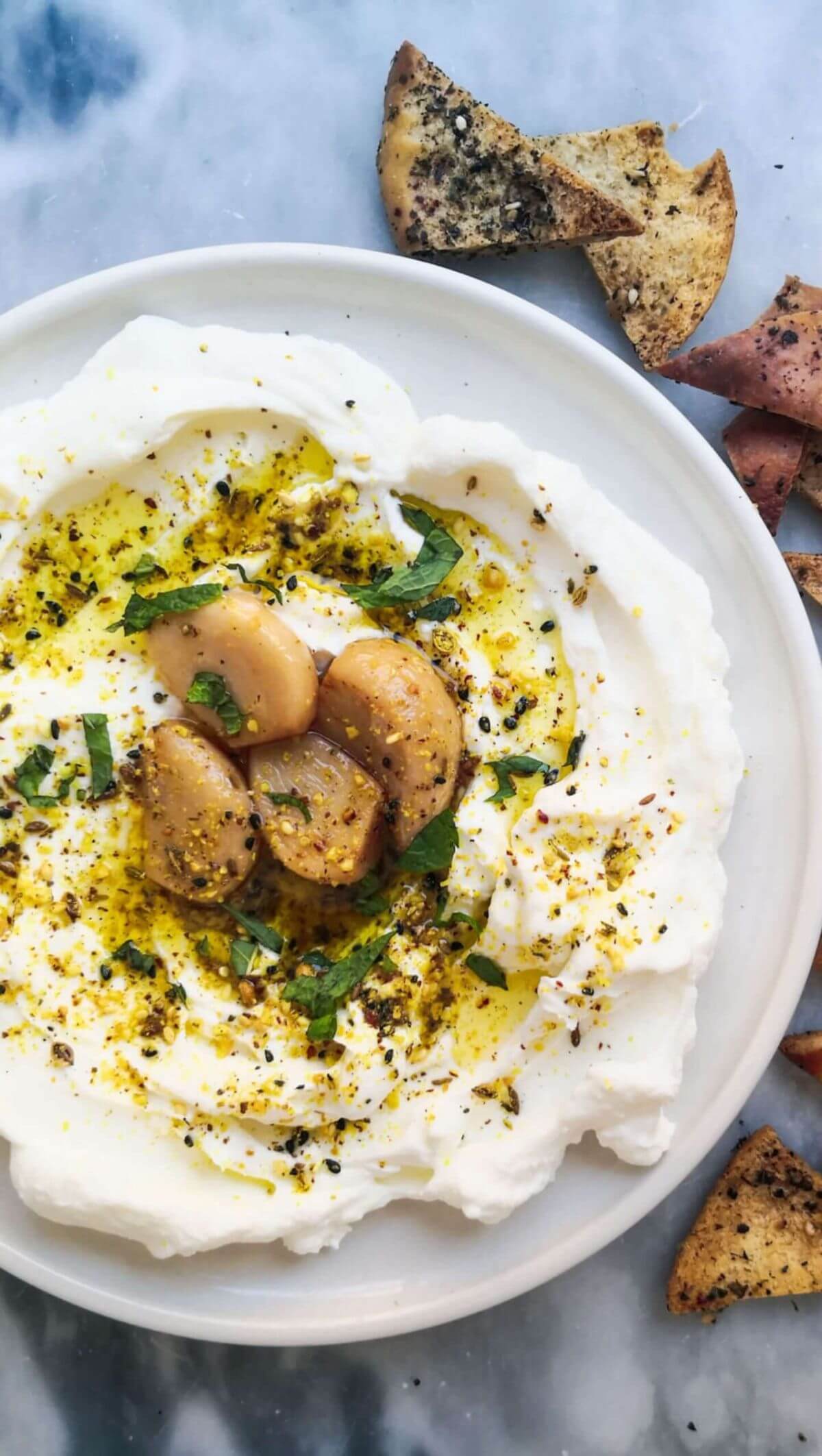 Labneh with confit garlic and dukkah on a plate with pita chips on the side.