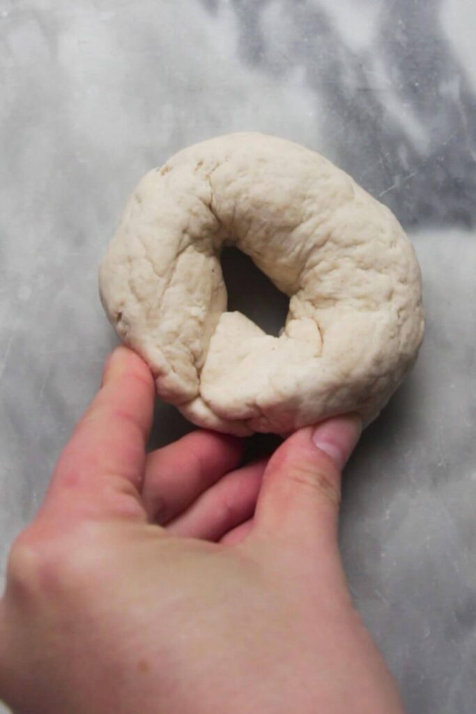 Hand shaping dough rope into a bagel on a grey marble surface.
