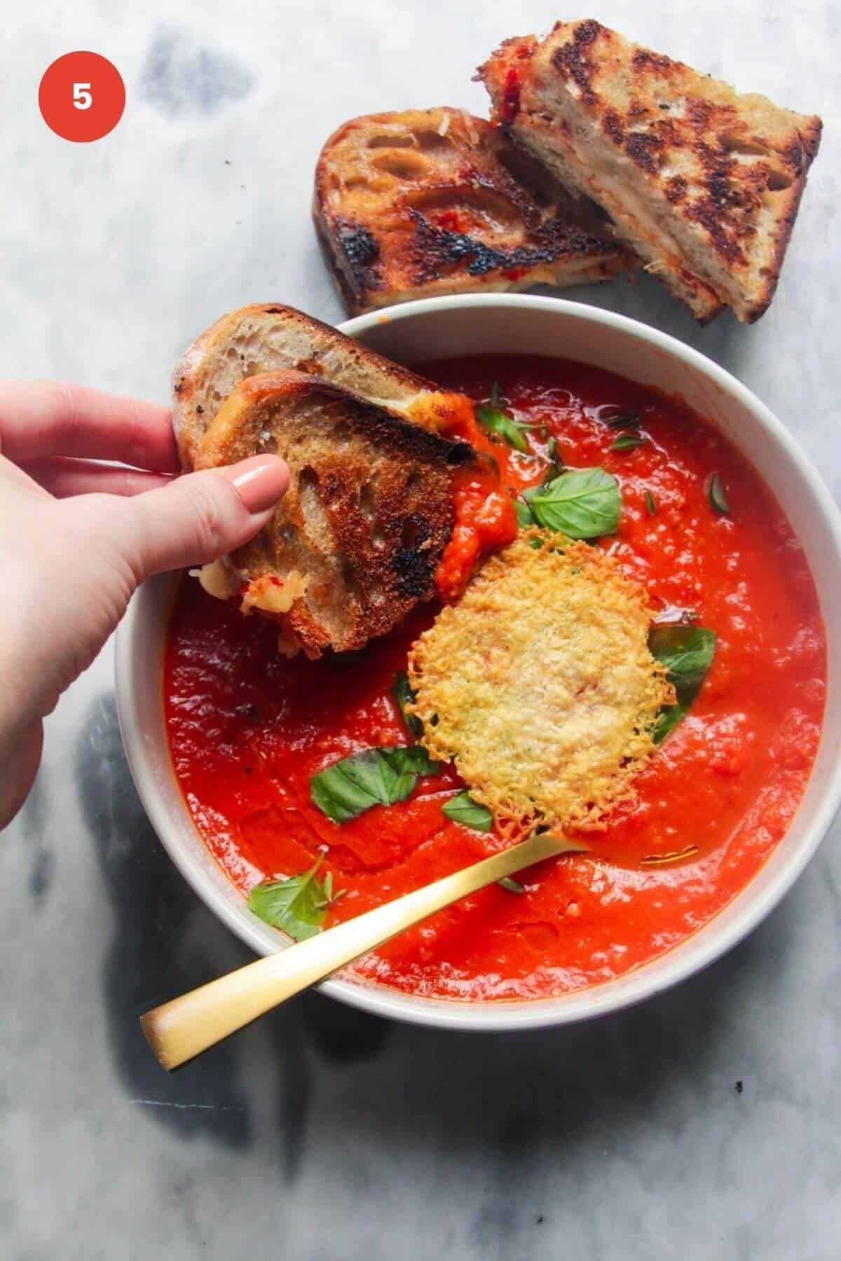 Hand dipping grilled cheese into tomato soup topped with parmesan crisp.