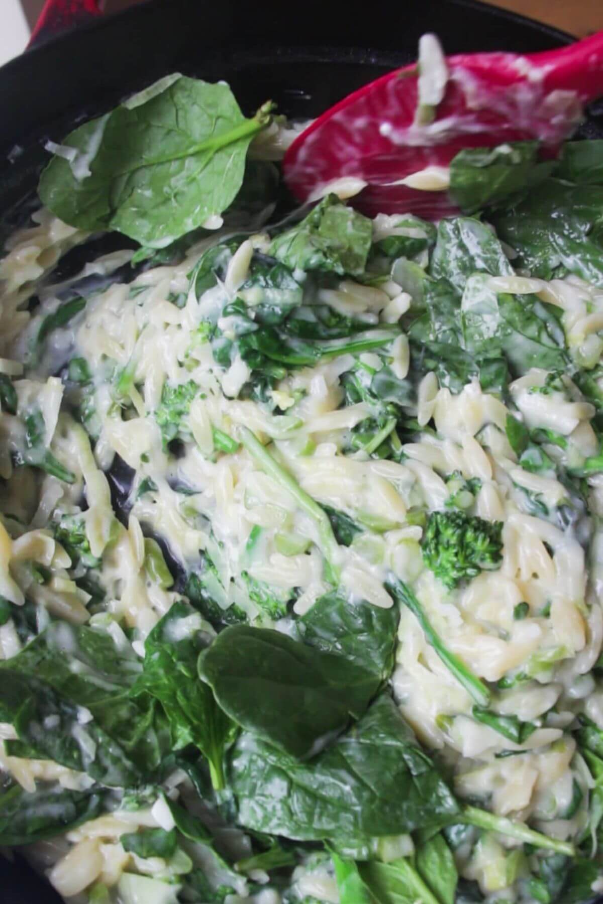 Spinach leaves being folded into creamy orzo with a red spatula.