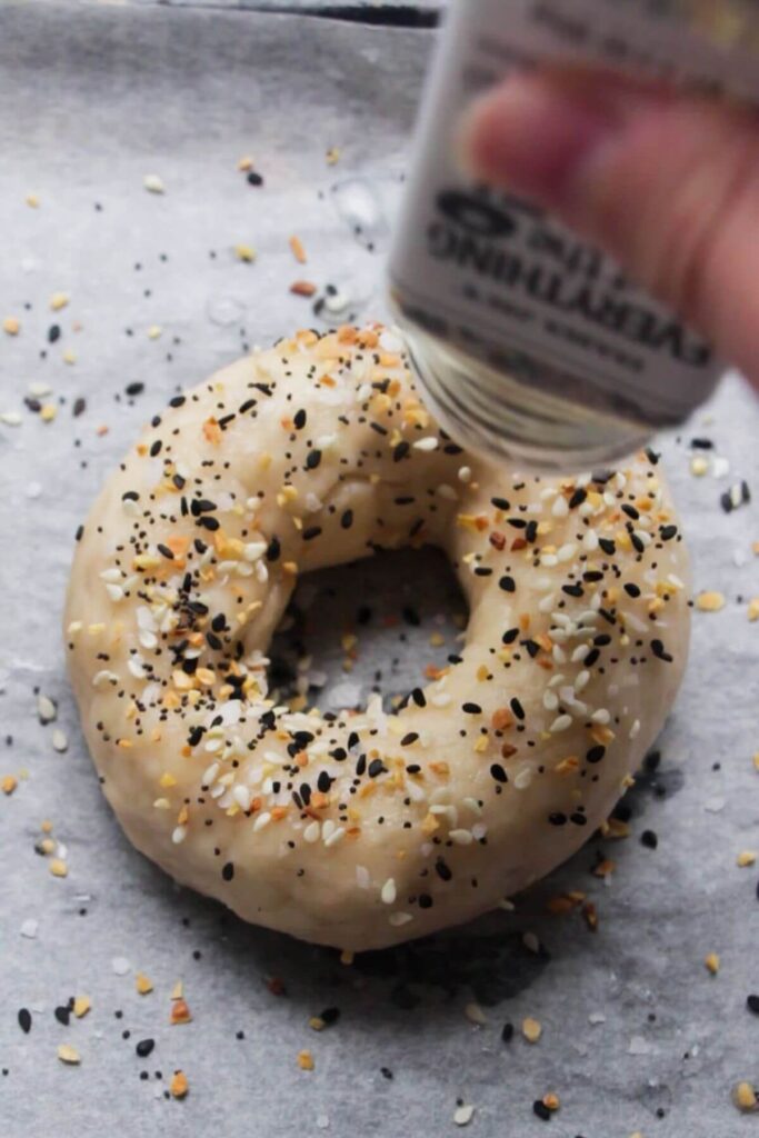 Everything bagel seasoning being sprinkled on top of bagels on a baking paper lined tray.