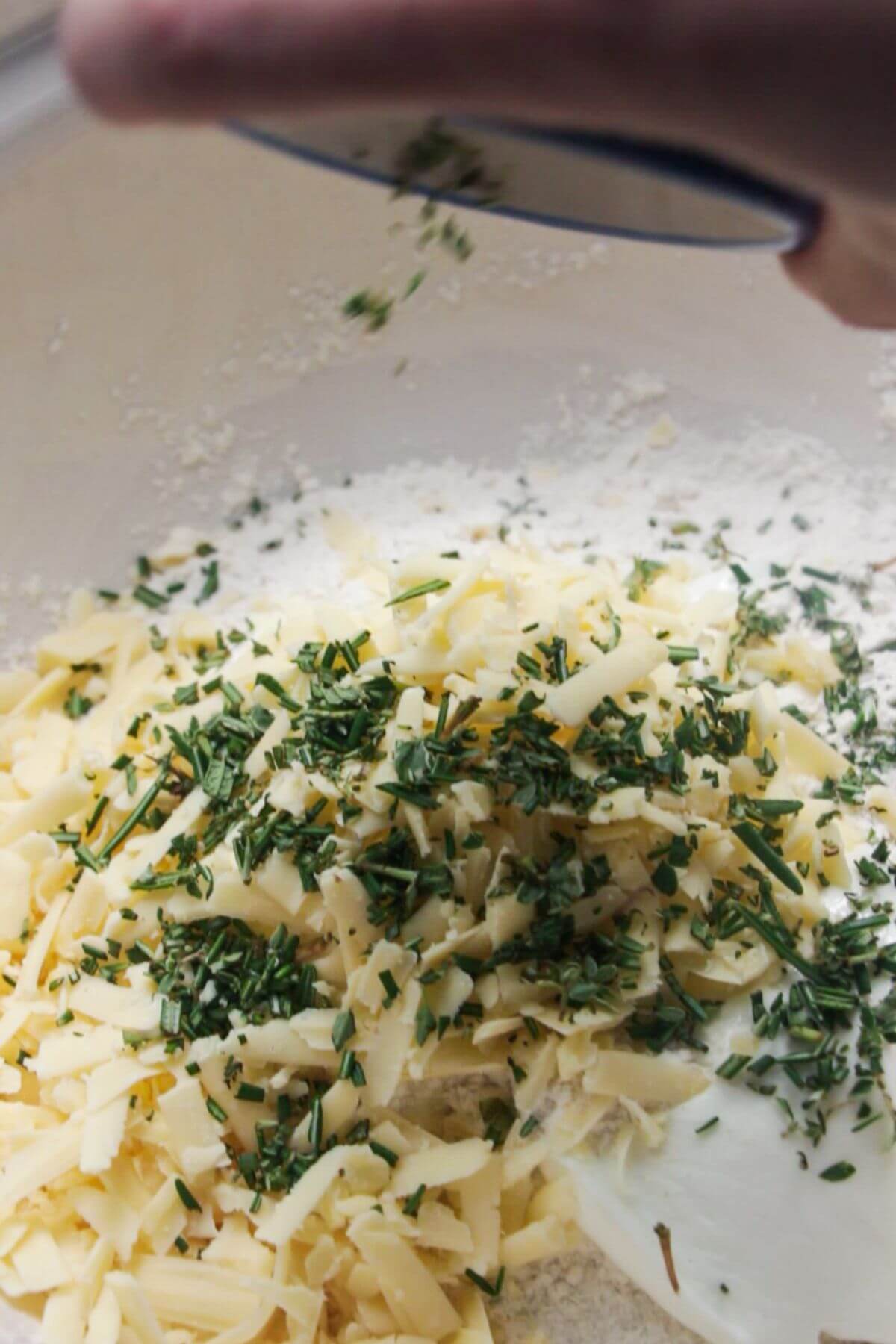 Chopped rosemary and thyme being added to a large bowl with yogurt, cheese and flour.