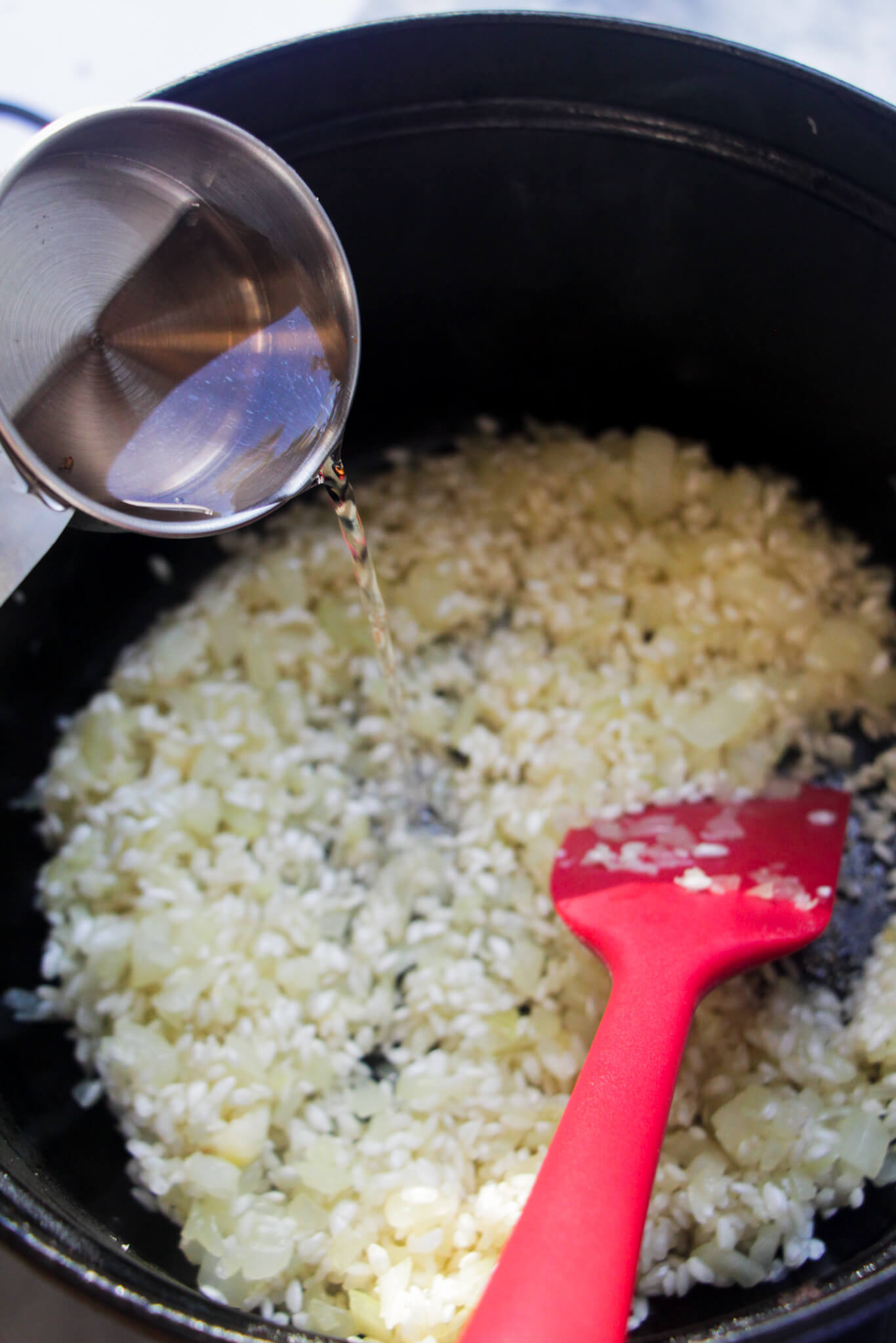 Silver cup measure adding white wine to rice, onion and garlic in a black pot with a red spatula in it.