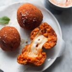 3 whole arancini on a small plate with a open arancini with cheese spilling out, with aioli with pesto on the side in a small bowl.