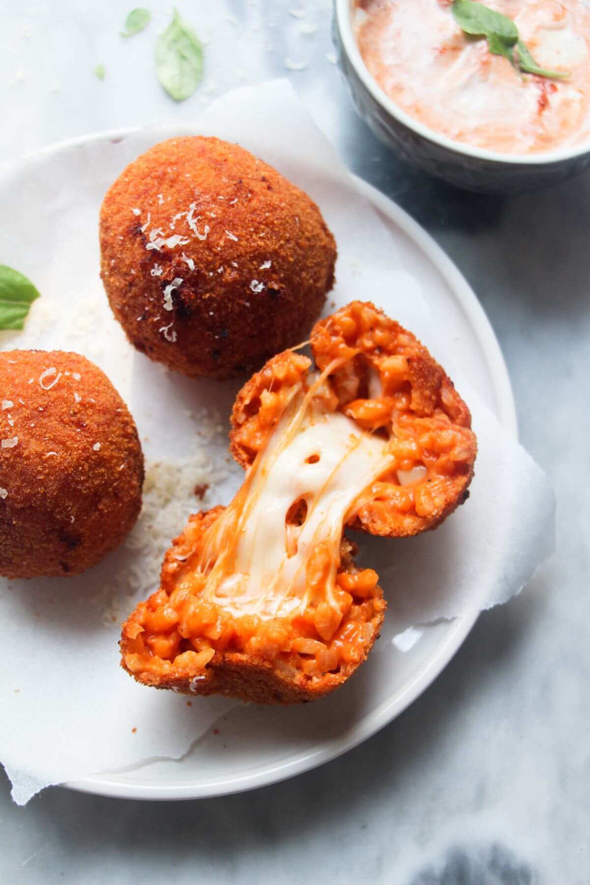 3 whole arancini on a small plate with a open arancini with cheese spilling out, with aioli with pesto on the side in a small bowl.