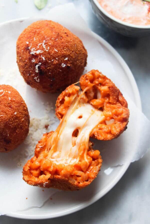 3 whole arancini on a small plate with a open arancini with cheese spilling out, with aioli with pesto on the side in a small bowl.