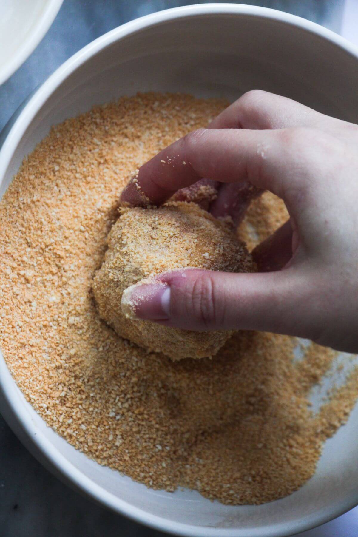 Hand tossing arancini in breadcrumbs in a white bowl.