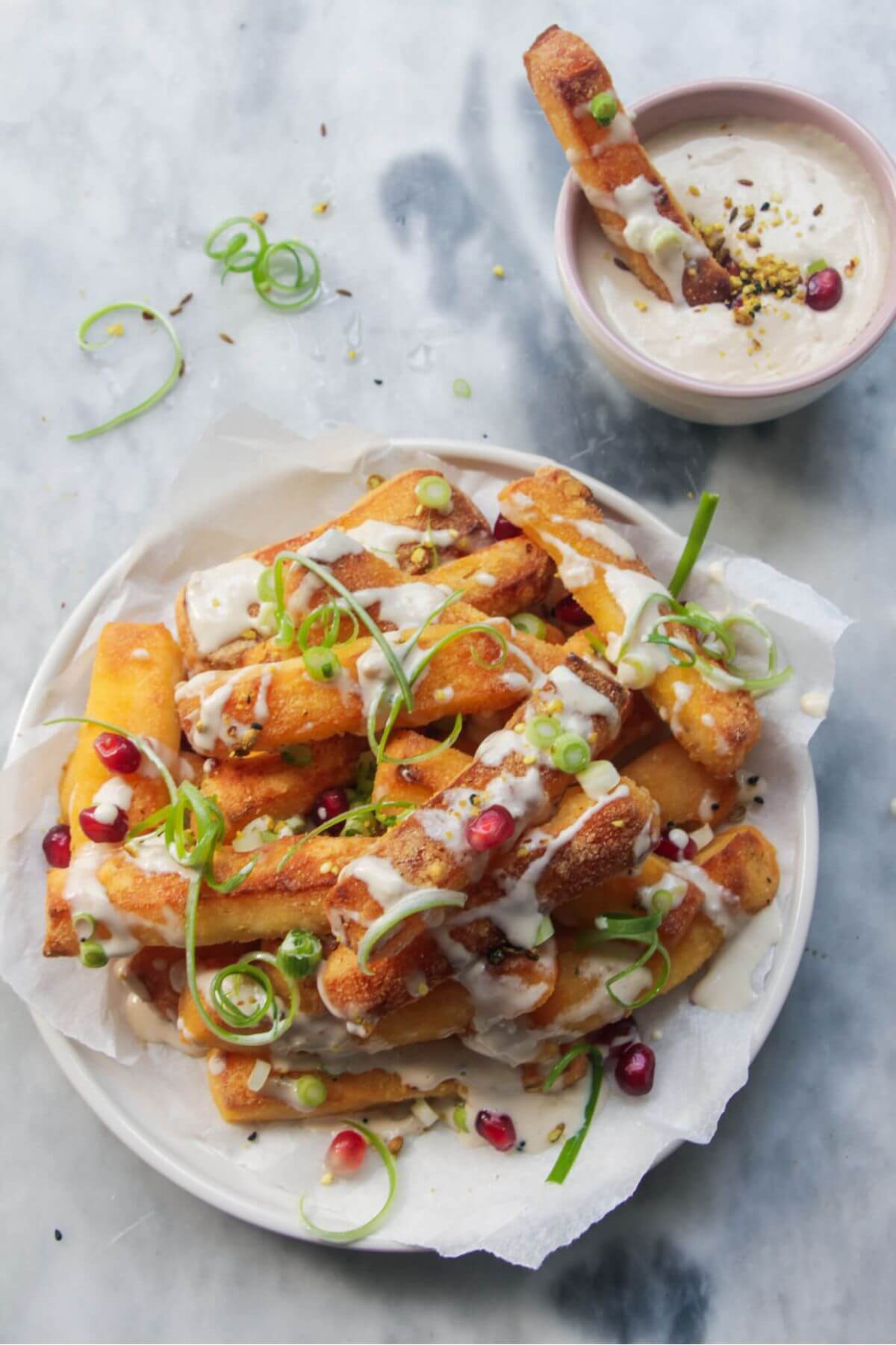 Pile of halloumi fries on a white plate with pomegranate seeds, spring onion and tahini sauce drizzled on top.