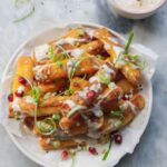Crispy halloumi fries piled onto a small white plate and drizzled with tahini sauce, spring onion and pomegranate seeds.