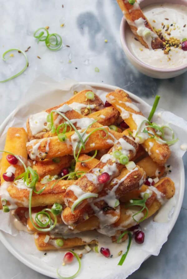 Crispy halloumi fries piled onto a small white plate and drizzled with tahini sauce, spring onion and pomegranate seeds with fry in a small bowl of tahini sauce on the side.