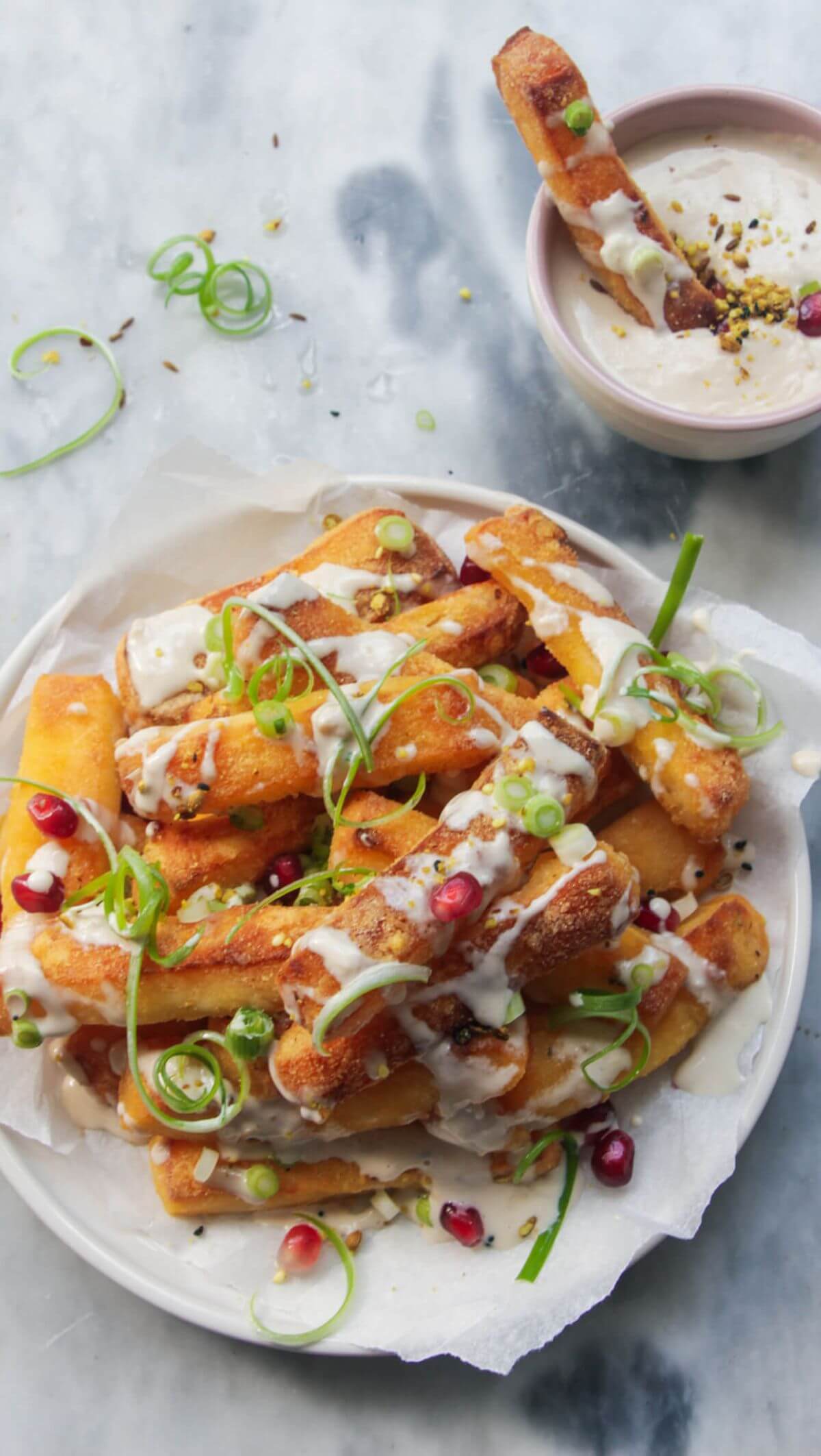 Crispy halloumi fries piled onto a small white plate and drizzled with tahini sauce, spring onion and pomegranate seeds with fry in a small bowl of tahini sauce on the side.
