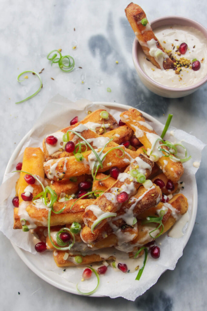 Crispy halloumi fries piled onto a small white plate and drizzled with tahini sauce, spring onion and pomegranate seeds.