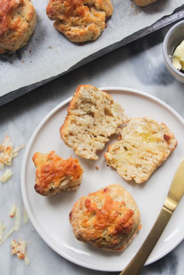 Cut open cheese scone on a small white plate, with whole scone next to it and more scones on oven tray in the background.