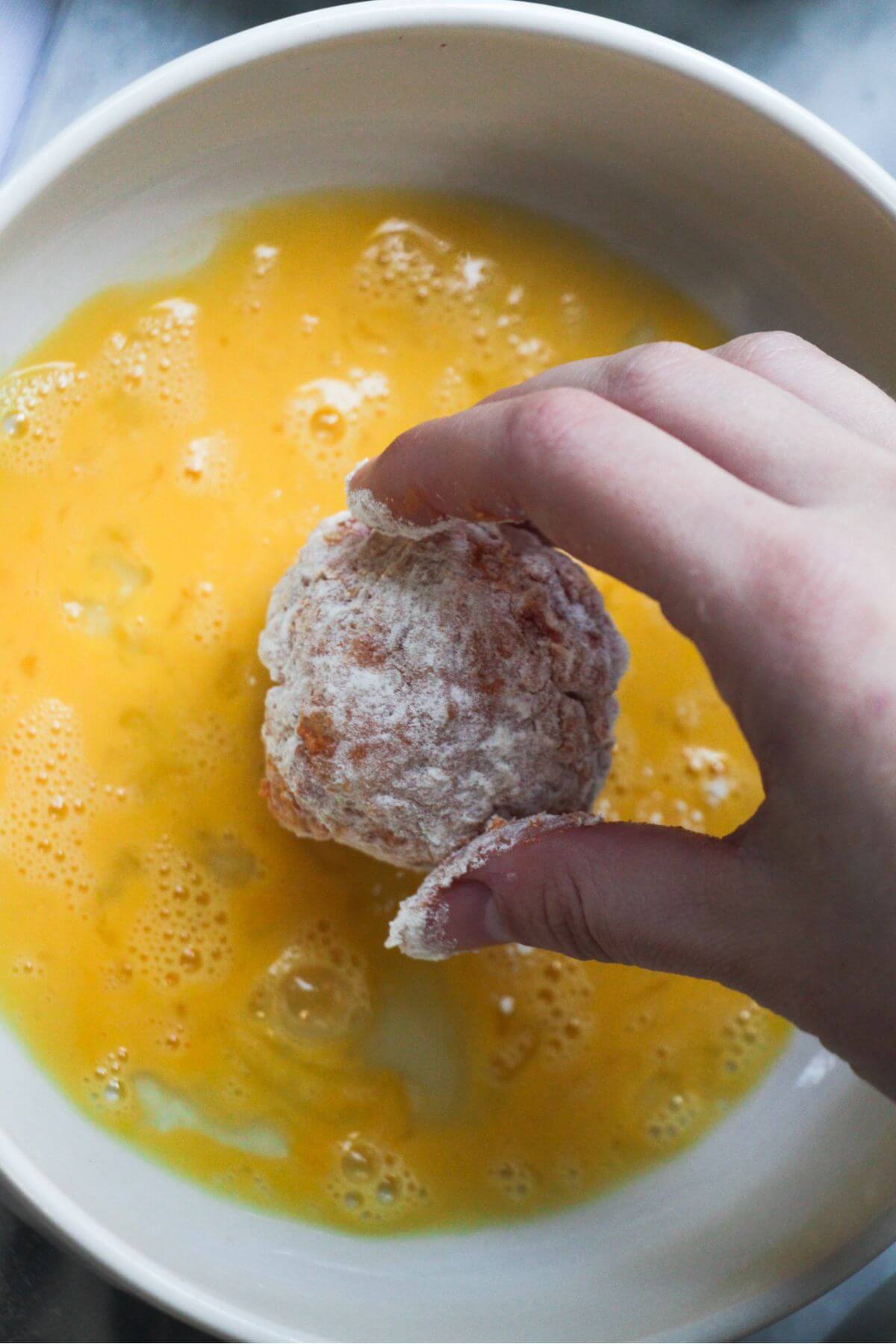 Hand placing flour coated arancini ball into a small bowl of whisked egg.